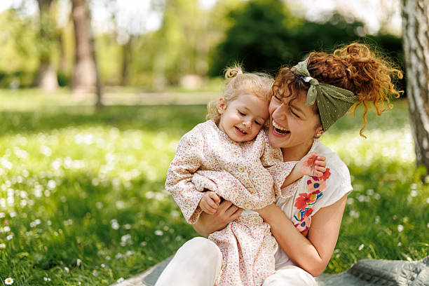 Mother and young daughter share a joyful hug on a sunny park lawn.